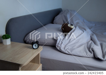 There is a mini alarm clock and a white potted plant on the bedside table, and a brown teddy bear is lying on a gray bed under a blanket in the background. The concept of a sleep mode, a comfortable There is a mini alarm clock and a white potted plant on the bedside table, and a brown teddy bear is lying on a gray bed under a blanket in the background. The concept of a sleep mode, a comfortable 142415405