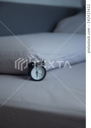 On the light gray bed linen there is a black miniature alarm clock against the background of two soft pillows in the dim lighting. Concept: the feeling of early morning, calm bedroom interior. High 142415412