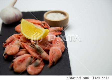 There is a handful of frozen shrimp with a slice of lemon on a black stone board, garlic and a salt shaker on the blurred background on the left. Concept: laconic serving of cold appetizers, emphasis There is a handful of frozen shrimp with a slice of lemon on a black stone board, garlic and a salt shaker on the blurred background on the left. Concept: laconic serving of cold appetizers, emphasis 142415415