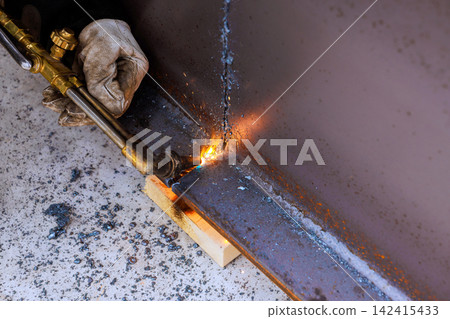 Worker focuses on cutting metal piece with using torch while standing in workshop. Worker focuses on cutting metal piece with using torch while standing in workshop. 142415433
