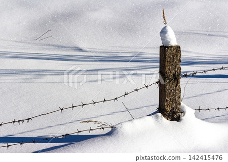 Gritty concrete fence post with barbed wire cuts across a vast snowy winter field. Sharp shadows stretch, emphasizing stark isolation and a defined perimeter Gritty concrete fence post with barbed wire cuts across a vast snowy winter field. Sharp shadows stretch, emphasizing stark isolation and a defined perimeter 142415476