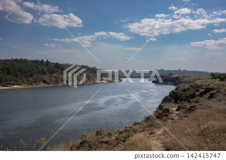 Wide view of a calm river flowing between rugged rocky banks under a bright blue sky with scattered clouds, capturing peaceful summer nature, scenic water and wild landscape beauty Wide view of a calm river flowing between rugged rocky banks under a bright blue sky with scattered clouds, capturing peaceful summer nature, scenic water and wild landscape beauty 142415747