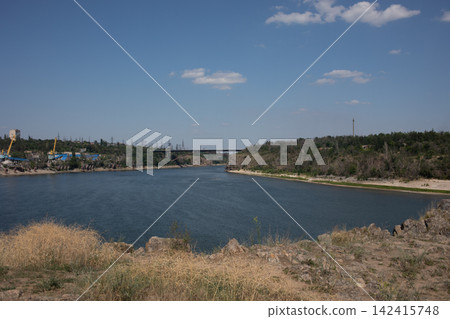 Wide view of a calm river bordered by dry grassy banks and green woodland beneath a clear blue sky, capturing peaceful summer scenery, natural landscape and tranquil freshwater nature Wide view of a calm river bordered by dry grassy banks and green woodland beneath a clear blue sky, capturing peaceful summer scenery, natural landscape and tranquil freshwater nature 142415748