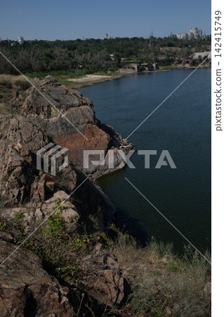Vertical view of rugged rocky cliffs beside calm blue river water, with sparse vegetation along the shore, capturing wild summer nature, dramatic terrain and peaceful landscape scenery Vertical view of rugged rocky cliffs beside calm blue river water, with sparse vegetation along the shore, capturing wild summer nature, dramatic terrain and peaceful landscape scenery 142415749