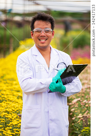 Smiling scientist man holding clipboard in flower nursery, wearing gloves and goggles for horticulture science research among colorful blooming plants Smiling scientist man holding clipboard in flower nursery, wearing gloves and goggles for horticulture science research among colorful blooming plants 142417133