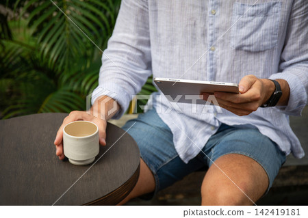 A freelancer relaxes with coffee, focused on a tablet outside A freelancer relaxes with coffee, focused on a tablet outside 142419181