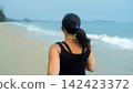 Rear view of young athletic woman in black outfit running beside calm sea with soft waves and overcast sky. Outdoor training concept 142423372
