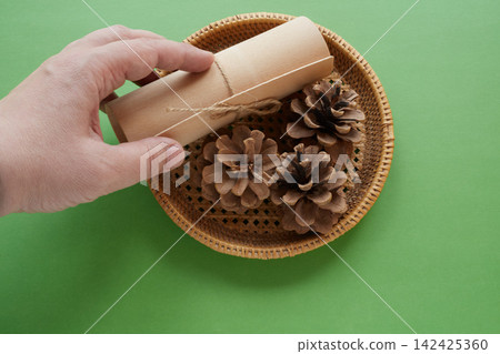 A hand is putting a gift wrap into a wicker basket with pine cones, green background, close-up.  142425360