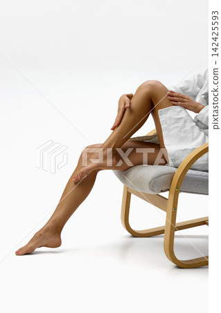 Woman touches smooth legs while relaxing in white bathrobe 142425593