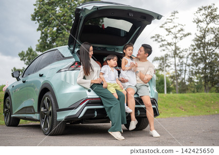 Asian family sharing a joyful conversation while sitting in an open SUV trunk in the green park. 142425650