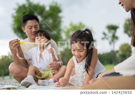 Asian father and son blowing with bubbles while daughter drawing with mother during a family picnic. Asian father and son blowing with bubbles while daughter drawing with mother during a family picnic. 142425658