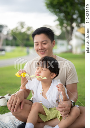 Asian father looking at son playing with bubbles blow stick while sitting on picnic blanket in park. 142425659