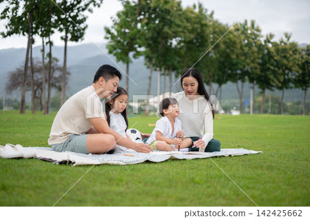 Asian family of four relaxing together on a picnic blanket mat surrounded by green park scenery. 142425662