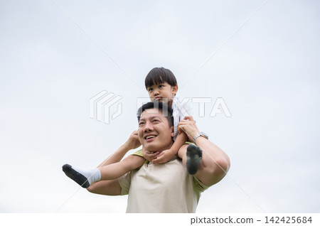 Asian father smiling while carrying a young kid son on his shoulders with a clear sky in background. 142425684