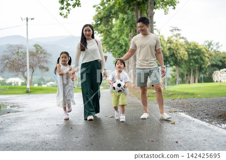 Asian family of parents and children walking together with a soccer ball on wet pathway in park. Asian family of parents and children walking together with a soccer ball on wet pathway in park. 142425695