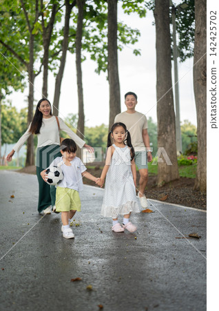 Asian children walking together with a soccer football while parents follow behind on park pathway. 142425702