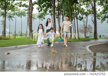 Asian parents walking together while watching their children running on a wet pathway in the park. Asian parents walking together while watching their children running on a wet pathway in the park. 142425703