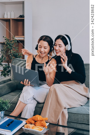 Two young female students using a laptop while sitting on a sofa at home. Student life concept with studying, teamwork, and online learning. Two young female students using a laptop while sitting on a sofa at home. Student life concept with studying, teamwork, and online learning. 142428682