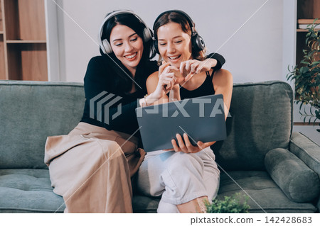 Two young female students using a laptop while sitting on a sofa at home. Student life concept with studying, teamwork, and online learning. Two young female students using a laptop while sitting on a sofa at home. Student life concept with studying, teamwork, and online learning. 142428683