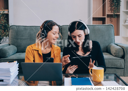 Two young female students using a laptop while sitting on a sofa at home. Student life concept with studying, teamwork, and online learning. Two young female students using a laptop while sitting on a sofa at home. Student life concept with studying, teamwork, and online learning. 142428708
