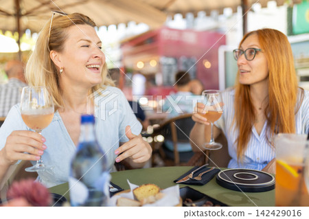 Two friends enjoy drinks and food at an outdoor cafe in a busy urban setting on a sunny day 142429016