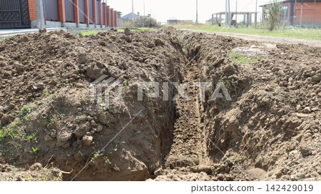 A long, deep trench dug in the ground along a suburban property. Along the edges lie piles of earth, and in the background, residential buildings and fences are visible on a warm, sunny day. 142429019