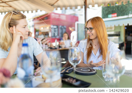 Two women talk at a cafe in an outdoor setting during a sunny day Two women talk at a cafe in an outdoor setting during a sunny day 142429020