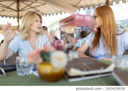 Two women having a conversation at a cafe during the day while sitting at a table Two women having a conversation at a cafe during the day while sitting at a table 142429036