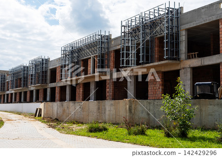 Row of unfinished concrete townhouses under construction with exposed steel reinforcement and brick infill along dusty access road, with construction, townhous, unfinished, concrete detail Row of unfinished concrete townhouses under construction with exposed steel reinforcement and brick infill along dusty access road, with construction, townhous, unfinished, concrete detail 142429296