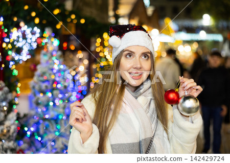 Girl in red Santa hat and with Christmas tree toys in her hands smiles dreamily Girl in red Santa hat and with Christmas tree toys in her hands smiles dreamily 142429724