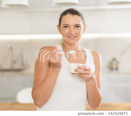 Adult woman eating fresh yogurt with spoon in modern kitchen Adult woman eating fresh yogurt with spoon in modern kitchen 142429871