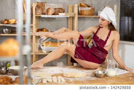 Naked woman in apron and chef hat sitting on flour-covered bakery table Naked woman in apron and chef hat sitting on flour-covered bakery table 142429888