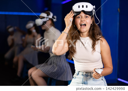 Girl gamer with virtual reality glasses on her forehead expresses enthusiastic emotions from playing with her friends in dark room Girl gamer with virtual reality glasses on her forehead expresses enthusiastic emotions from playing with her friends in dark room 142429998