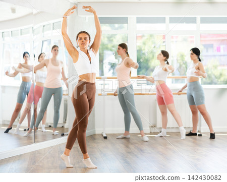 Young beautiful female ballet student demonstrating skills in ballet in dance studio. Group of young ballet dancers in background near ballet barre Young beautiful female ballet student demonstrating skills in ballet in dance studio. Group of young ballet dancers in background near ballet barre 142430082