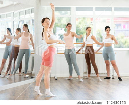 Young beautiful female ballet student demonstrating skills in ballet in dance studio. Group of young ballet dancers in background near ballet barre Young beautiful female ballet student demonstrating skills in ballet in dance studio. Group of young ballet dancers in background near ballet barre 142430153
