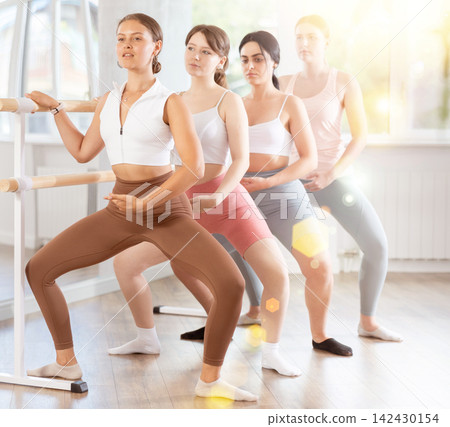 Group of sports young girls standing in row together while doing ballet dance moves demi plie while holding on ballet barre in ballet studio Group of sports young girls standing in row together while doing ballet dance moves demi plie while holding on ballet barre in ballet studio 142430154