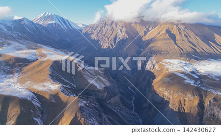 Aerial view of the stunning Caucasus mountain range in Georgia, showcasing rugged peaks, snow-capped summits, and deep valleys under a clear blue sky with scattered clouds Aerial view of the stunning Caucasus mountain range in Georgia, showcasing rugged peaks, snow-capped summits, and deep valleys under a clear blue sky with scattered clouds 142430627