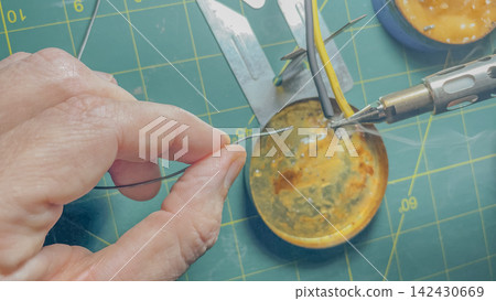Close-up of a hand holding a thin wire while soldering electronic components on a green cutting mat with various tools and a soldering iron visible in the background Close-up of a hand holding a thin wire while soldering electronic components on a green cutting mat with various tools and a soldering iron visible in the background 142430669