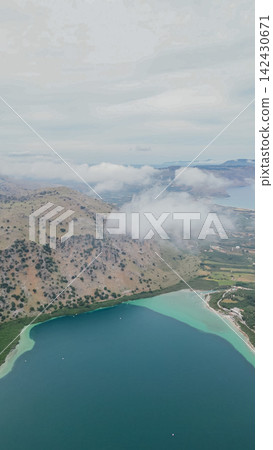 Aerial view of Lake Kournas in Crete, surrounded by green hills and clouds, showcasing the clear turquoise waters and diverse landscape of the region Aerial view of Lake Kournas in Crete, surrounded by green hills and clouds, showcasing the clear turquoise waters and diverse landscape of the region 142430671