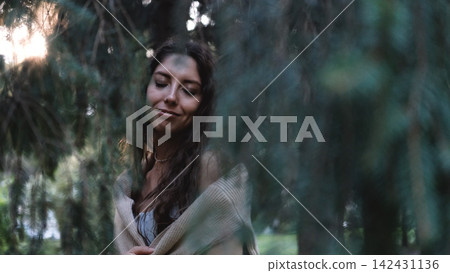 Young woman framed by hanging evergreen branches in forest, calm shifting gaze, soft backlight, cool green tones, textured knit clothing, and intimate vertical portrait mood with quiet woodland Young woman framed by hanging evergreen branches in forest, calm shifting gaze, soft backlight, cool green tones, textured knit clothing, and intimate vertical portrait mood with quiet woodland 142431136
