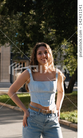 A portrait of a cheerful girl with long curly hair smiling at the camera. A close-up portrait of a friendly young woman in casual clothes against a blurred city background. Outdoors. A portrait of a cheerful girl with long curly hair smiling at the camera. A close-up portrait of a friendly young woman in casual clothes against a blurred city background. Outdoors. 142431141