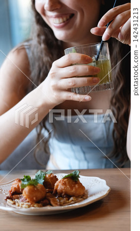 Young woman smiles while stirring pale drink in short glass at dining table with plated food in foreground, indoor close view with soft natural light, blurred background, warm wood surface, relaxed Young woman smiles while stirring pale drink in short glass at dining table with plated food in foreground, indoor close view with soft natural light, blurred background, warm wood surface, relaxed 142431213