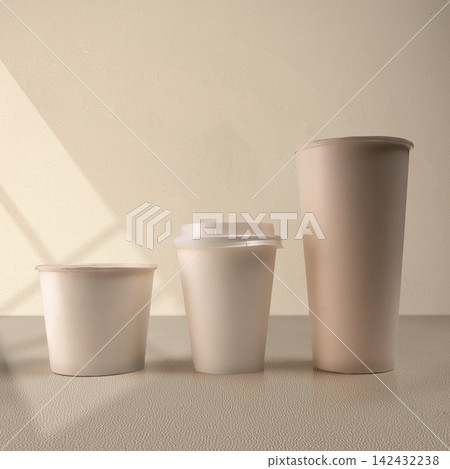 Three blank beige paper cups of different sizes lined up on a neutral surface with soft shadow overlay, perfect for packaging mockup. Mockup or Copyspace Three blank beige paper cups of different sizes lined up on a neutral surface with soft shadow overlay, perfect for packaging mockup. Mockup or Copyspace 142432238