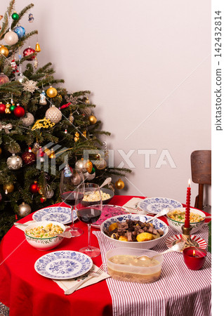 A table set for a Christmas and New Year celebration displays various dishes like salad, meat, and grains. A red candle, wine glass, and colorful decorations add to the festive atmosphere A table set for a Christmas and New Year celebration displays various dishes like salad, meat, and grains. A red candle, wine glass, and colorful decorations add to the festive atmosphere 142432814