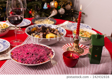 A table set for a Christmas and New Year celebration displays various dishes like salad, meat, and grains. A red candle, wine glass, and colorful decorations add to the festive atmosphere A table set for a Christmas and New Year celebration displays various dishes like salad, meat, and grains. A red candle, wine glass, and colorful decorations add to the festive atmosphere 142432816