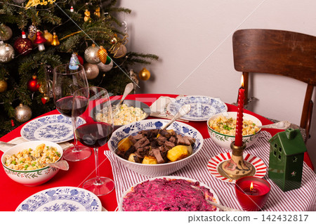 A table set for a Christmas and New Year celebration displays various dishes like salad, meat, and grains. A red candle, wine glass, and colorful decorations add to the festive atmosphere A table set for a Christmas and New Year celebration displays various dishes like salad, meat, and grains. A red candle, wine glass, and colorful decorations add to the festive atmosphere 142432817