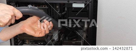 A 53-year-old man uses a vacuum attachment to remove dust from a computer system unit in a home office. The room is well lit and organized for the task, banner A 53-year-old man uses a vacuum attachment to remove dust from a computer system unit in a home office. The room is well lit and organized for the task, banner 142432871