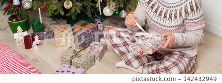 A woman aged 52 sits on the floor next to a decorated Christmas tree. She opens gifts on Christmas Day while enjoying the holiday with family members in the background A woman aged 52 sits on the floor next to a decorated Christmas tree. She opens gifts on Christmas Day while enjoying the holiday with family members in the background 142432949