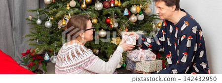 On Christmas Day, a 52-year-old couple spends time together near a decorated tree. They exchange gifts and celebrate the holiday in their cozy home setting filled with decorations On Christmas Day, a 52-year-old couple spends time together near a decorated tree. They exchange gifts and celebrate the holiday in their cozy home setting filled with decorations 142432950