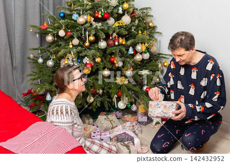 A 52-year-old couple sits on the floor, smiling and opening gifts. The room is decorated with a Christmas tree, colorful ornaments, and presents around them. It is Christmas Day A 52-year-old couple sits on the floor, smiling and opening gifts. The room is decorated with a Christmas tree, colorful ornaments, and presents around them. It is Christmas Day 142432952